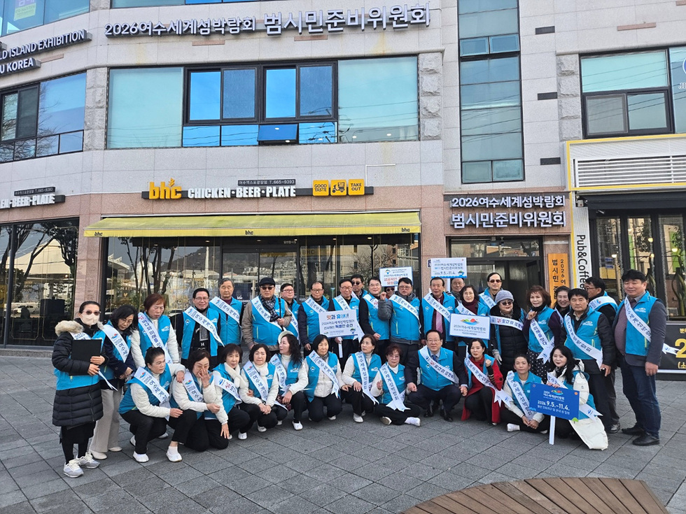 2026 여수 세계 섬 박람회 범시민 준비위원회가 bhc 치킨과 맥주 가게 앞에서 모여 사진을 찍고 있다. 위원들은 파란색 조끼를 입고 있으며, 여러 개의 현수막과 팻말을 들고 있다.
