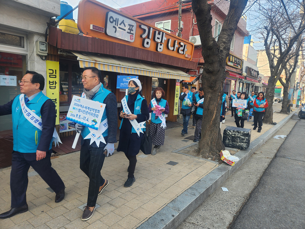 2026 여자 지방선거와 방사선 치료위원회를 알리는 행사가 진행되고 있으며, 참가자들이 거리에서 피켓을 들고 걷고 있다.
