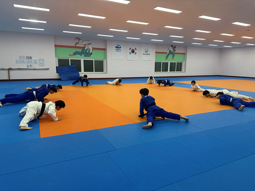 체육관에서 유도복을 입은 어린이들이 훈련 중인 모습이다. 바닥은 주황색과 파란색으로 나뉘어 있으며, 벽에는 유도 관련 그림이 있다.