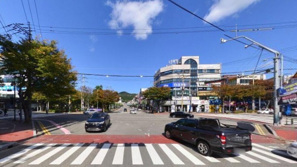 한적한 도로의 교차로에서 차량들이 지나가고 있으며, 주변에는 상점들이 늘어서 있다. 맑은 하늘 아래 가로수들이 보인다.