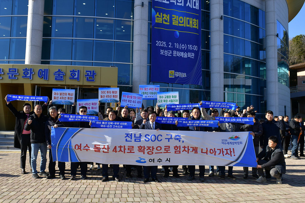 여수 돌산 4차로 확장을 촉구하는 집회가 문화예술회관 앞에서 열리고 있으며, 참가자들이 관련 현수막과 피켓을 들고 있다.