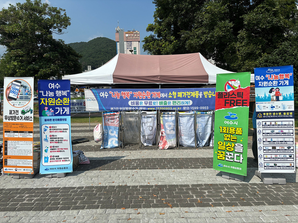 여수에서 열린 자원순환 가게에서 소형 폐기물품을 수거하는 행사 장면으로, 다양한 안내 현수막과 쓰레기 수거함이 배치되어 있다.