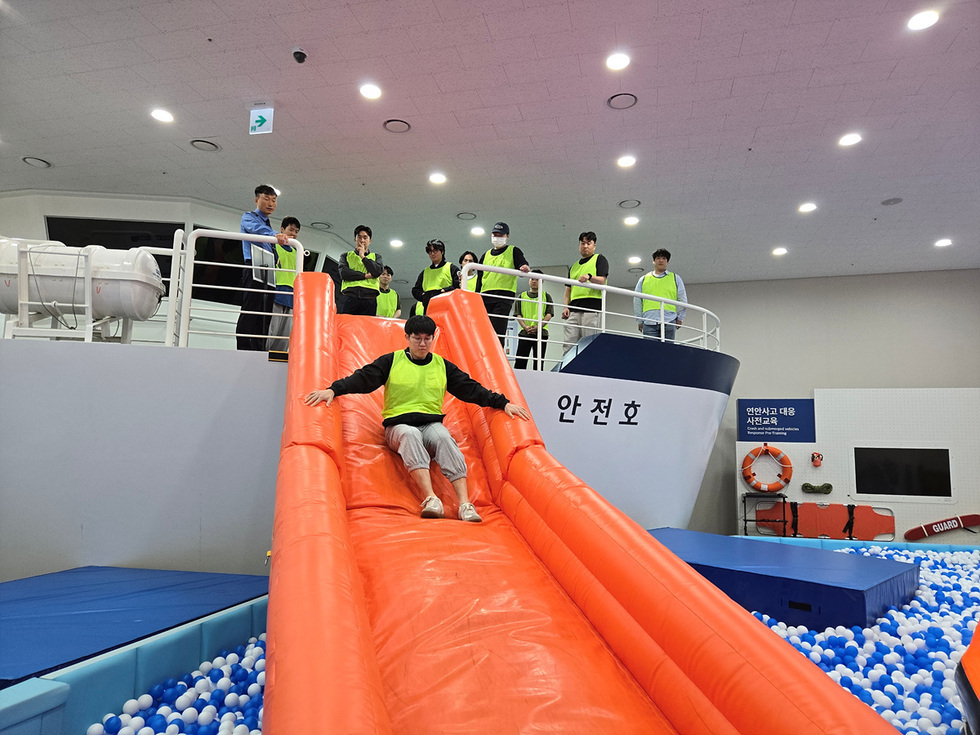 안전 교육을 받는 사람들이 배 모형 위에서 슬라이드를 타고 있는 장면이다. 한 남성이 슬라이드를 내려오고 있으며, 다른 사람들은 위에서 지켜보고 있다.