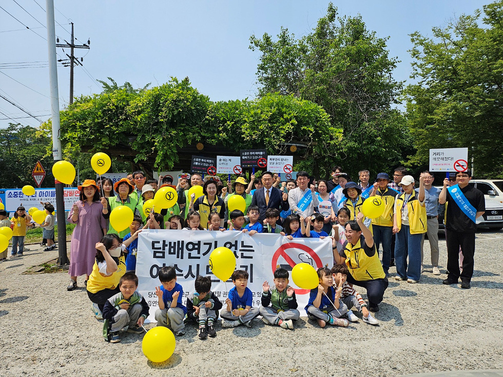 여수에서 담배연기 없는 환경을 위한 집회가 열리고 있으며, 참가자들이 노란 풍선을 들고 구호를 외치고 있다.