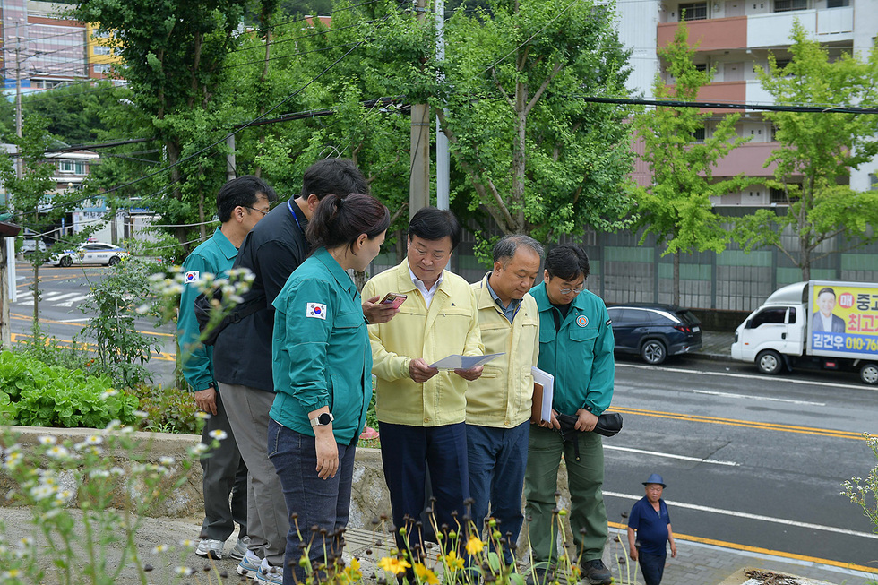 여러 명의 사람들이 서서 문서를 검토하고 있는 장면으로, 한 남성이 스마트폰을 사용하고 있다. 배경에는 중고차 광고가 보인다.