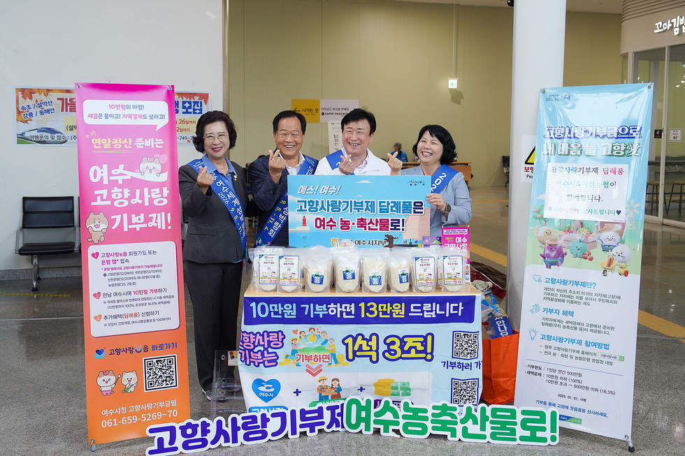 여수시청 관계자들이 고향사랑기부제를 홍보하며 귀성객들에게 쌀을 나누는 장면으로, 다양한 농산물과 기부 안내가 담긴 테이블이 배경에 있다.