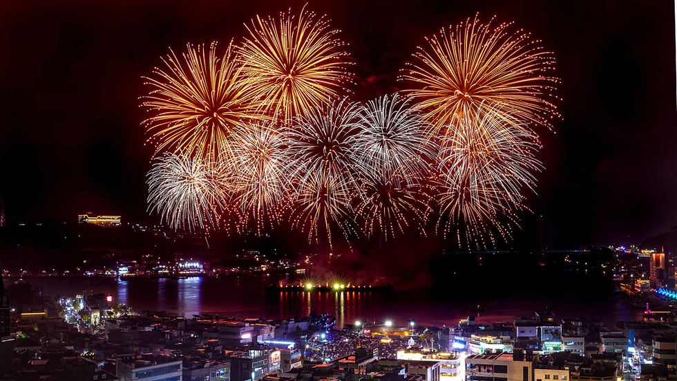 여수 도심과 바다 위에서 밤하늘을 수놓는 대형 불꽃이 연이어 터지는 장면. 불꽃이 반짝이며 바다와 도심 건물, 교량의 불빛이 어우러져 환상적인 야경을 이루고 있다