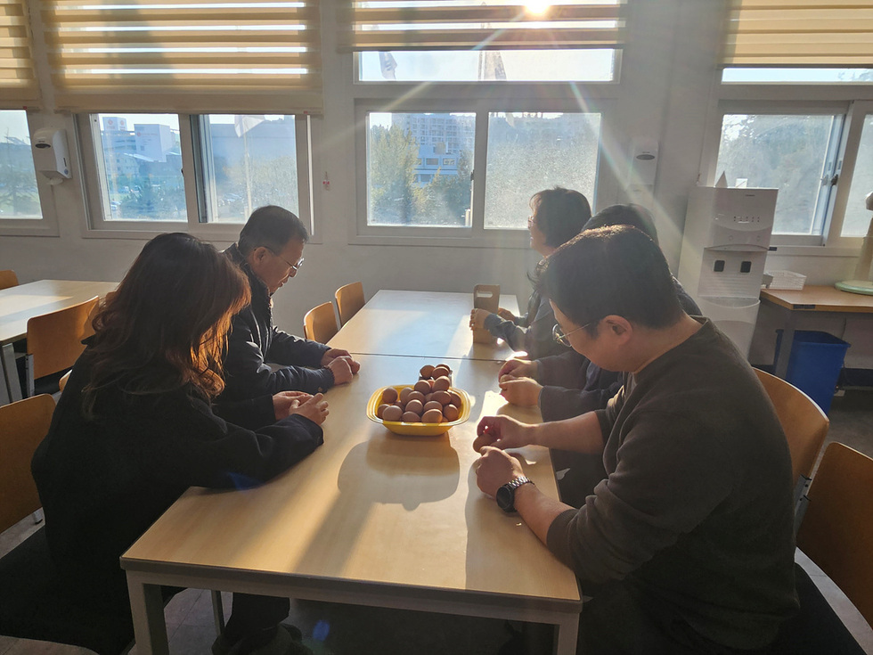 창가에서 햇빛이 들어오는 식당에서 직원들이 테이블에 둘러앉아 노란 접시에 담긴 구운계란을 놓고 함께 대화를 나누는 모습. 창밖으로 건물과 나무가 보이며 따뜻한 아침 분위기가 느껴진다