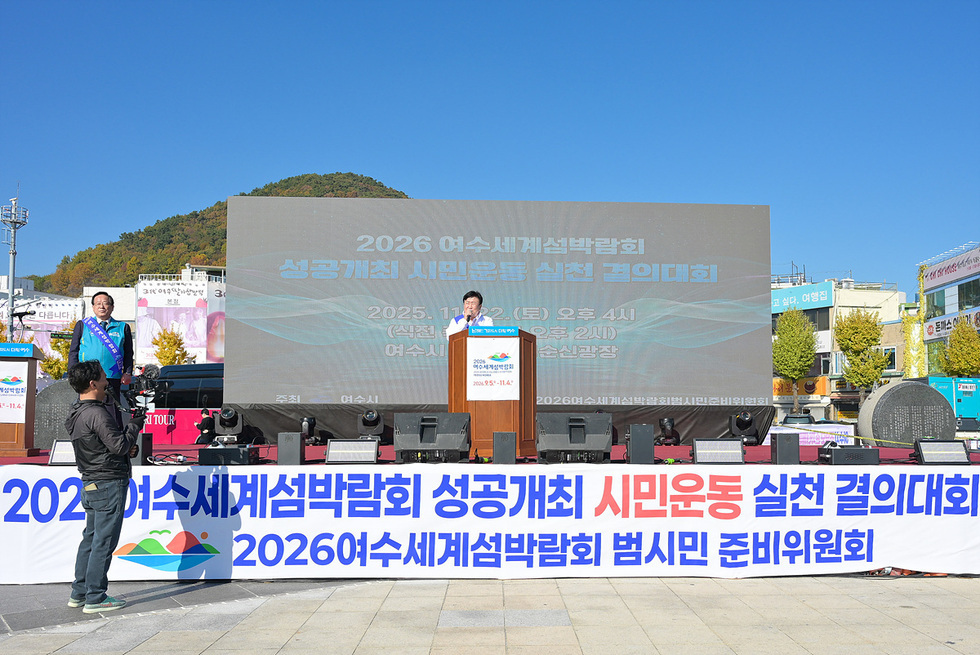 이순신광장 야외무대에서 열린 ‘2026여수세계섬박람회 성공개최 시민운동 실천 결의대회’ 현장. 큰 전광판 앞에서 연단에 선 발표자가 연설을 하고 있으며, 무대 아래에는 ‘2026여수세계섬박람회 성공개최 시민운동 실천 결의대회’라고 적힌 긴 현수막이 설치되어 있다.
