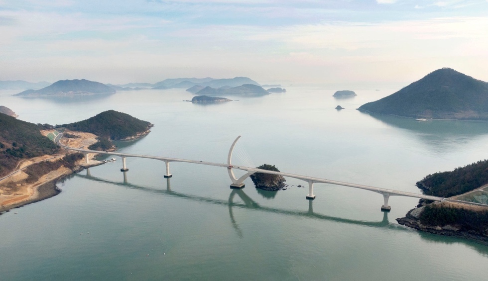 여수와 고흥을 잇는 해상교량이 잔잔한 바다 위에 길게 놓여 있는 모습. 주변에 크고 작은 섬들이 펼쳐져 있으며, 아치형 구조물이 있는 교량이 맑은 수면에 반사되어 보인다.