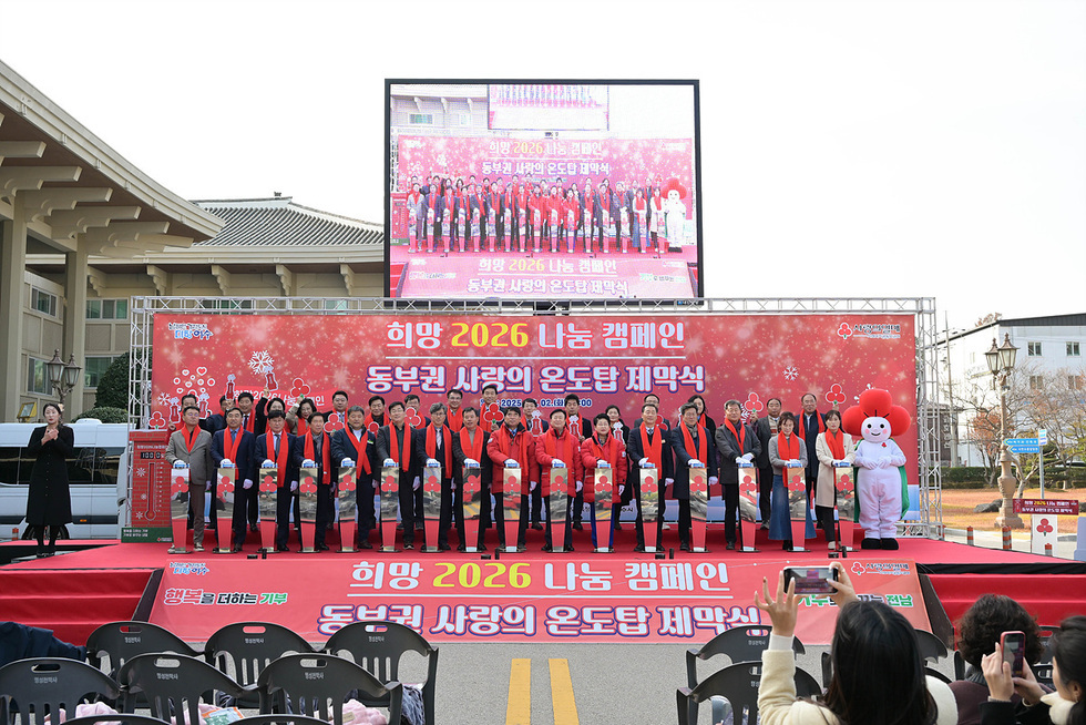 여수시청 앞에서 열린 희망2026 나눔캠페인 사랑의 온도탑 제막식 행사에서 정기명 여수시장과 참석자들이 무대 위에서 붉은 목도리를 두르고 손잡이를 누르는 세레모니를 진행하고 있으며 뒤편 대형 스크린에는 행사 모습이 비쳐지고 있다.