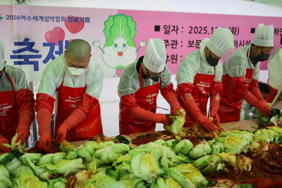 여수시 자비나눔 김장김치 대축제 행사장에서 참여자들이 빨간 앞치마와 위생모, 장갑을 착용한 채 배추에 양념을 버무려 김장을 담그고 있다. 작업대 위에는 절인 배추와 고춧가루 양념이 가득 펼쳐져 있으며, 뒤편 현수막에는 ‘2026여수세계섬박람회 성공기원’이 적혀 있다.